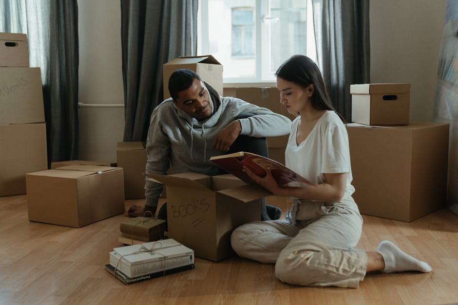 Inside a home during a house removal process, a young woman with shoulder-length dark hair, wearing a white top, is seated on the floor and organizing packing materials. In the background, ordely stac