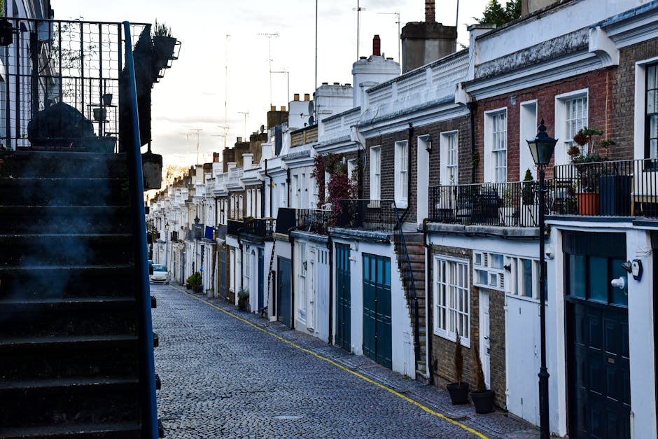 A narrow residential street with a cobblestone pavement during daylight, lined with terraced brick houses featuring white and blue exterior doors, small front gardens with potted plants, and black wrought iron railings. Some houses have external staircases leading to upper floors. On the left side, a black metal staircase with wooden steps is partially visible, possibly leading to an upper-level entrance. The street is quiet, with a few parked cars visible further down, and the sky above is overcast. This scene illustrates a typical urban neighbourhood where house removals and furniture transport by Removal Van Chiswick often require careful navigation of tight spaces, especially on narrow streets like this one, which are common in residential areas involved in home relocation services.