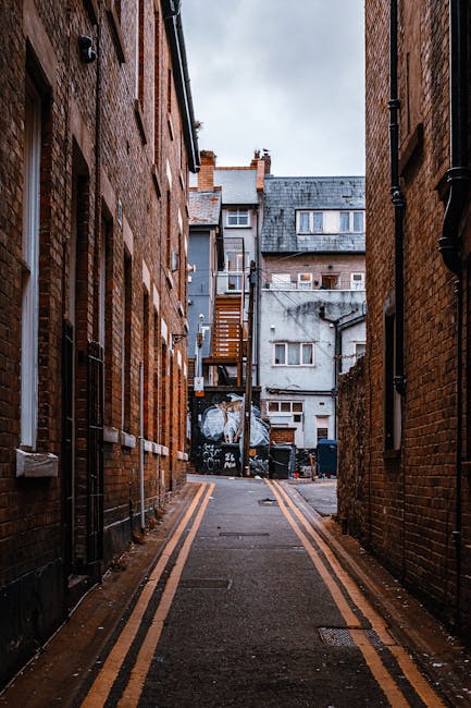 A narrow urban alleyway between two brick buildings during daytime, with double yellow lines painted on the road surface. At the far end, there are additional residential buildings, some with external staircases and small windows. The alley contains parked bins and waste containers near a graffiti-covered wall, and is illuminated by natural light from the overcast sky above. The scene depicts typical elements of a city street environment suitable for house removals and furniture transport, highlighting the tight space constraints encountered during home relocation projects, as handled by Removal Van Chiswick, who specialise in removals and moving logistics in difficult street conditions.