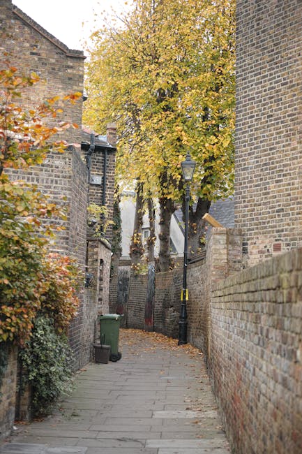A narrow residential street with a cobblestone pavement during daylight, lined with terraced brick houses featuring white and blue exterior doors, small front gardens with potted plants, and black wrought iron railings. Some houses have external staircases leading to upper floors. On the left side, a black metal staircase with wooden steps is partially visible, possibly leading to an upper-level entrance. The street is quiet, with a few parked cars visible further down, and the sky above is overcast. This scene illustrates a typical urban neighbourhood where house removals and furniture transport by Removal Van Chiswick often require careful navigation of tight spaces, especially on narrow streets like this one, which are common in residential areas involved in home relocation services.