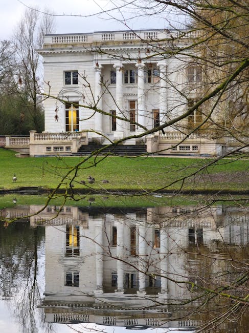A white, multi-story historic house with classical architectural features including tall windows, columns, and a balustrade on the roof terrace, situated on a grassy bank beside a calm river that reflects the building’s facade. In the foreground, leafless tree branches partially obscure the view, indicating a winter or early spring setting. The area around the house is landscaped with a low stone wall and steps leading to the entrance. This scene exemplifies the type of property involved in home relocation or furniture transport services, as offered by Removal Van Chiswick, with potential for loading or packing processes within the property visible in related context.