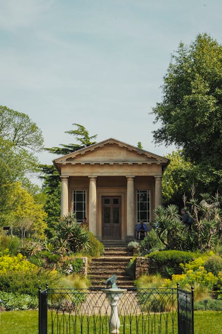 A historic stone building with classical architectural features, including four columns supporting a triangular pediment, is situated within a lush garden surrounded by a variety of green plants and trees. The structure is accessed by a short staircase leading to a wooden door, flanked by tall windows on each side. The garden in front is enclosed by a black metal fence and features a stone pathway, decorative plants, and a small fountain. The scene is taken during daylight with clear skies and bright natural light, emphasizing the peaceful, well-maintained environment indicative of a residential or estate setting suitable for house removals or relocations, as often managed by professional removal companies like Removal Van Chiswick.