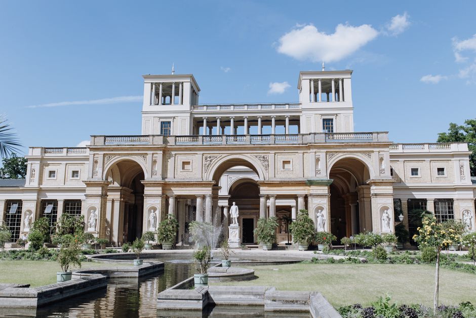A white, multi-story historic house with classical architectural features including tall windows, columns, and a balustrade on the roof terrace, situated on a grassy bank beside a calm river that reflects the building’s facade. In the foreground, leafless tree branches partially obscure the view, indicating a winter or early spring setting. The area around the house is landscaped with a low stone wall and steps leading to the entrance. This scene exemplifies the type of property involved in home relocation or furniture transport services, as offered by Removal Van Chiswick, with potential for loading or packing processes within the property visible in related context.
