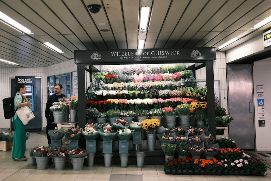 Inside a subway station, a flower stall operated by Wheeler of Chiswick displays a variety of colourful flowers arranged in metal buckets and packaging materials. The stall is situated against a tiled